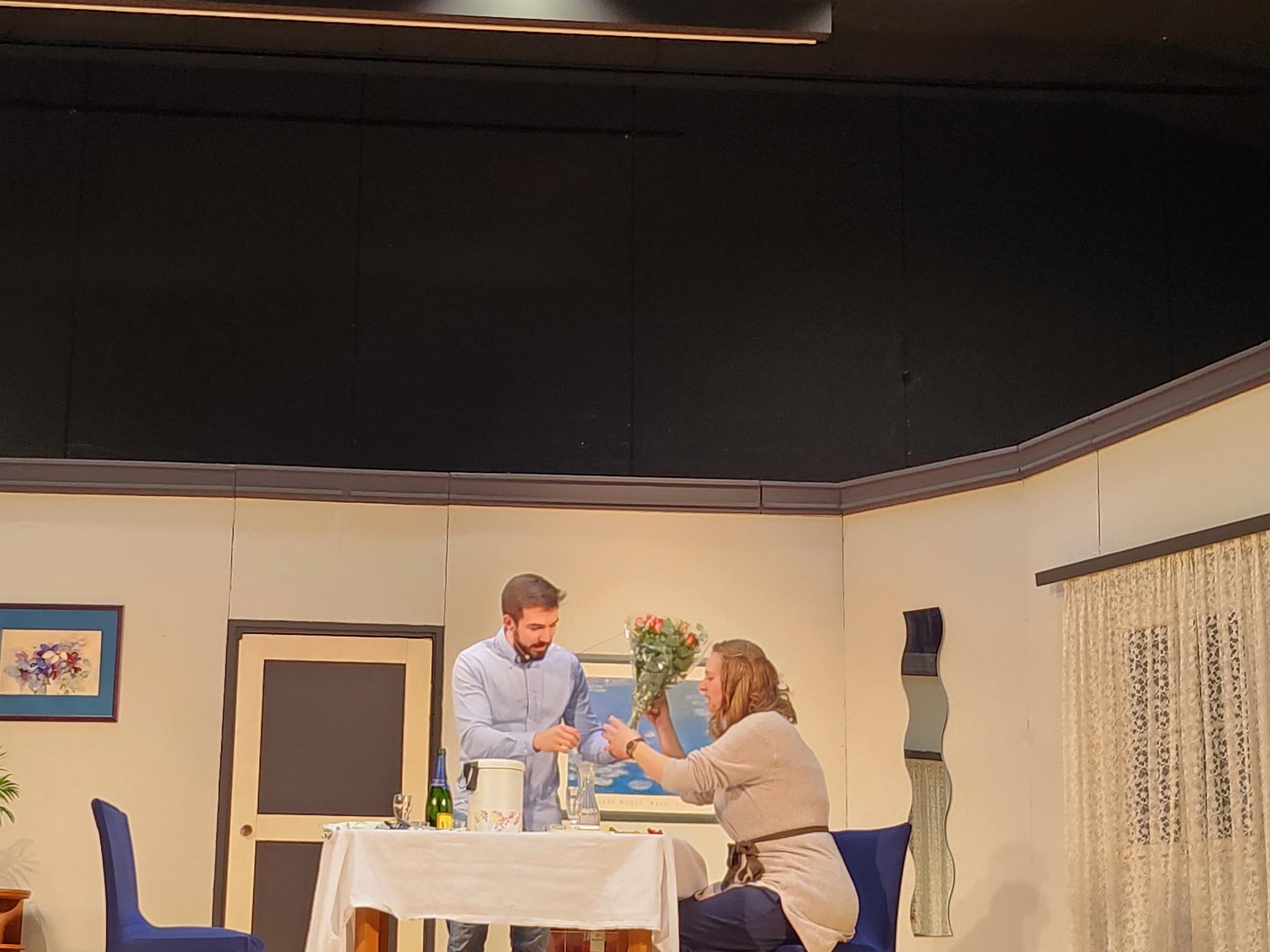Ein stehender Mann und eine sitzende Frau an einem gedeckten Tisch auf der Bühne, im Hintergrund die Theaterkulisse die ein Zimmer darstellt