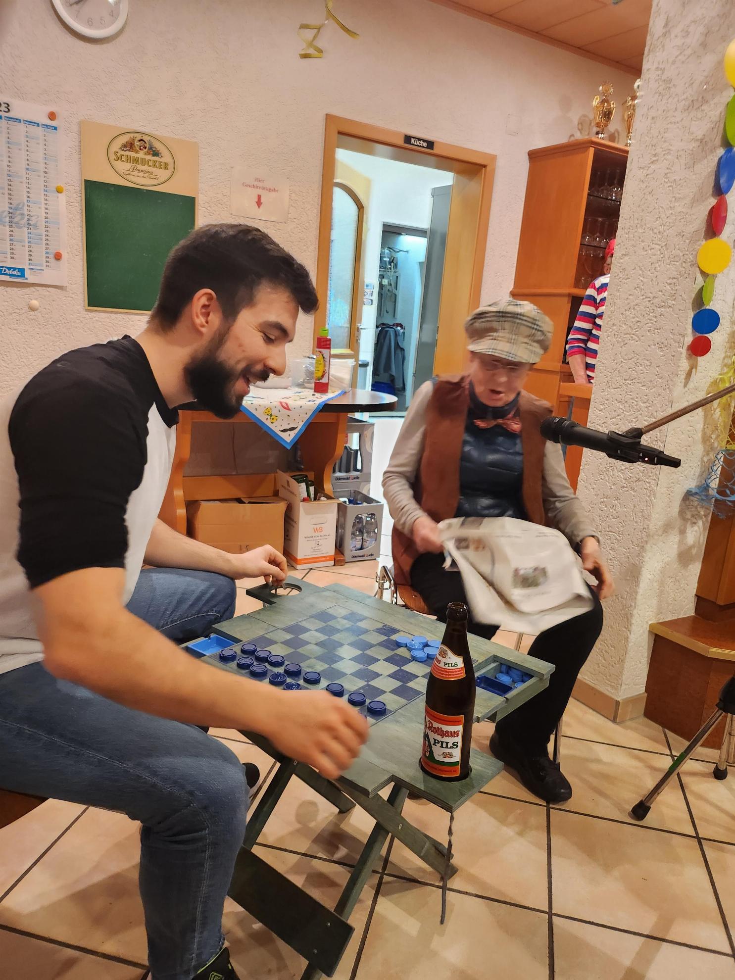 Ein junger Mann unterhällt sich mit einer älterne Frau, sie sitzen dabei an einem Tisch mit Schachbrettmuster auf dem eine Bierflasche steht