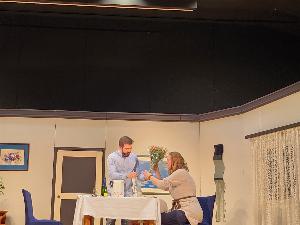 Ein stehender Mann und eine sitzende Frau an einem gedeckten Tisch auf der Bühne, im Hintergrund die Theaterkulisse die ein Zimmer darstellt