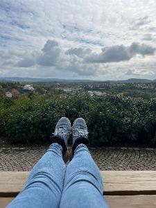 Ausgestreckte Beine, die in Richtung einer grünen, hügeligen Landschaft ragen. Weiße Wolken am Himmel.