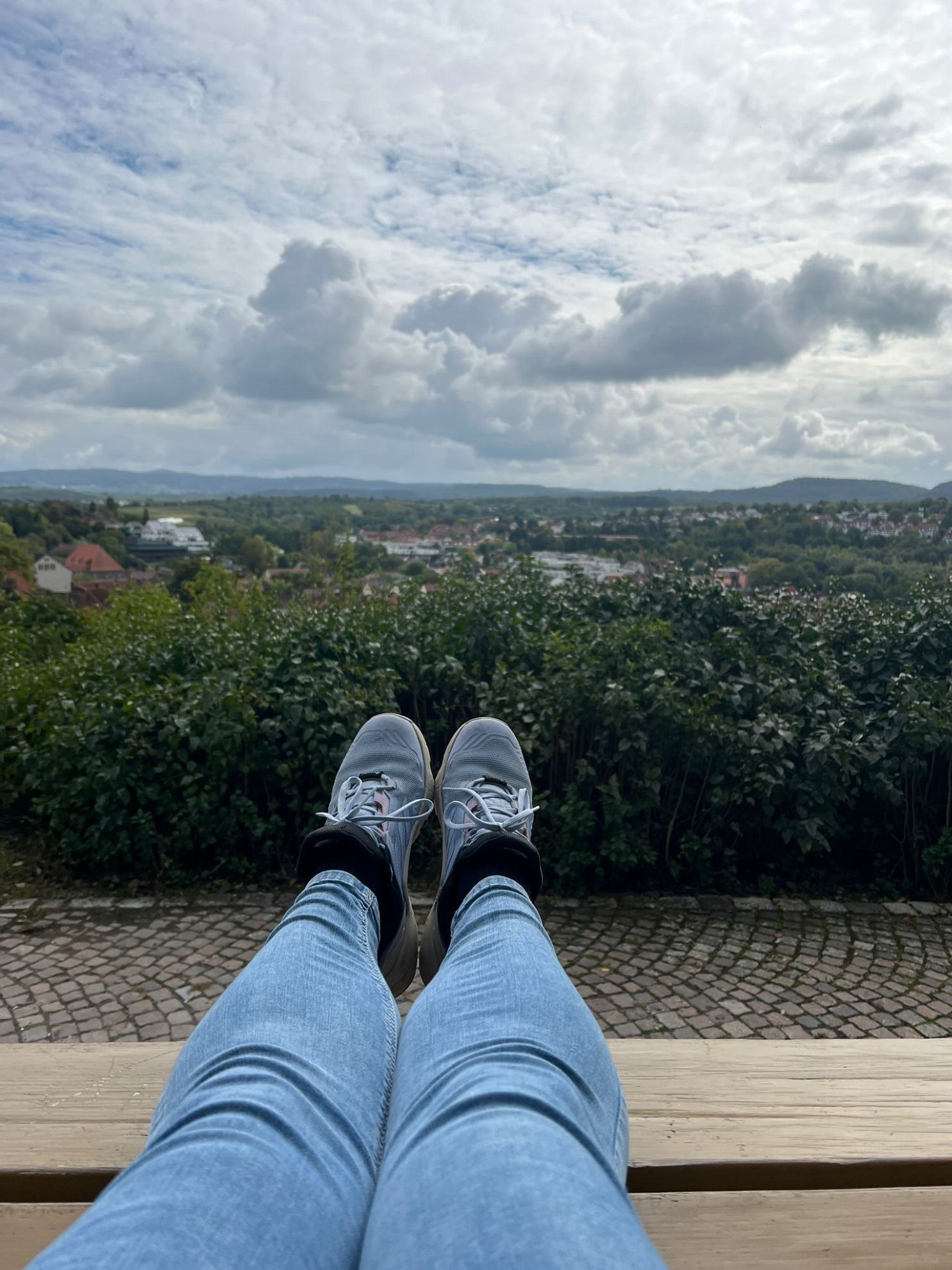 Ausgestreckte Beine, die in Richtung einer grünen, hügeligen Landschaft ragen. Weiße Wolken am Himmel.