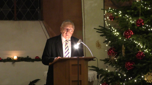 Ein Mann steht an einem Rednerpult, neben ihm ein festlich geschmückter Weihnachtsbaum.