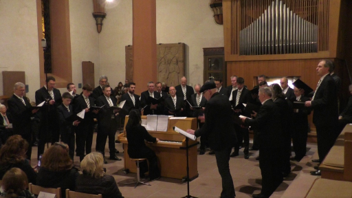 Ein Männerchor singt in der evangelischen Kirche in Schönau. Der Dirigent steht dabei neben einem Klavier zum Publikum gewandt und singt einen Solopart. Die Pianistin begleitet den Chor.