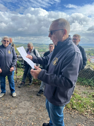 Jemand steht im Vordergund, ein Notenblatt in der Hand und singt unter blauem Himmel mit weißen Wolken. Im Hintergrund stehen Leute.